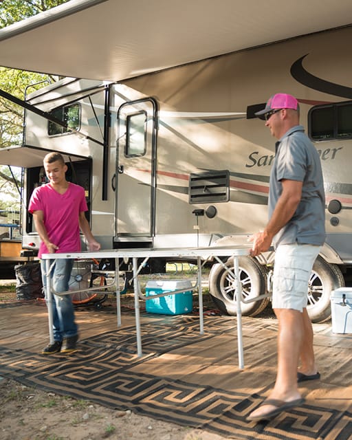 Three people set up an outdoor space next to a large camper. One person holds a folding table, another assembles it, while a third person exits the camper carrying items.