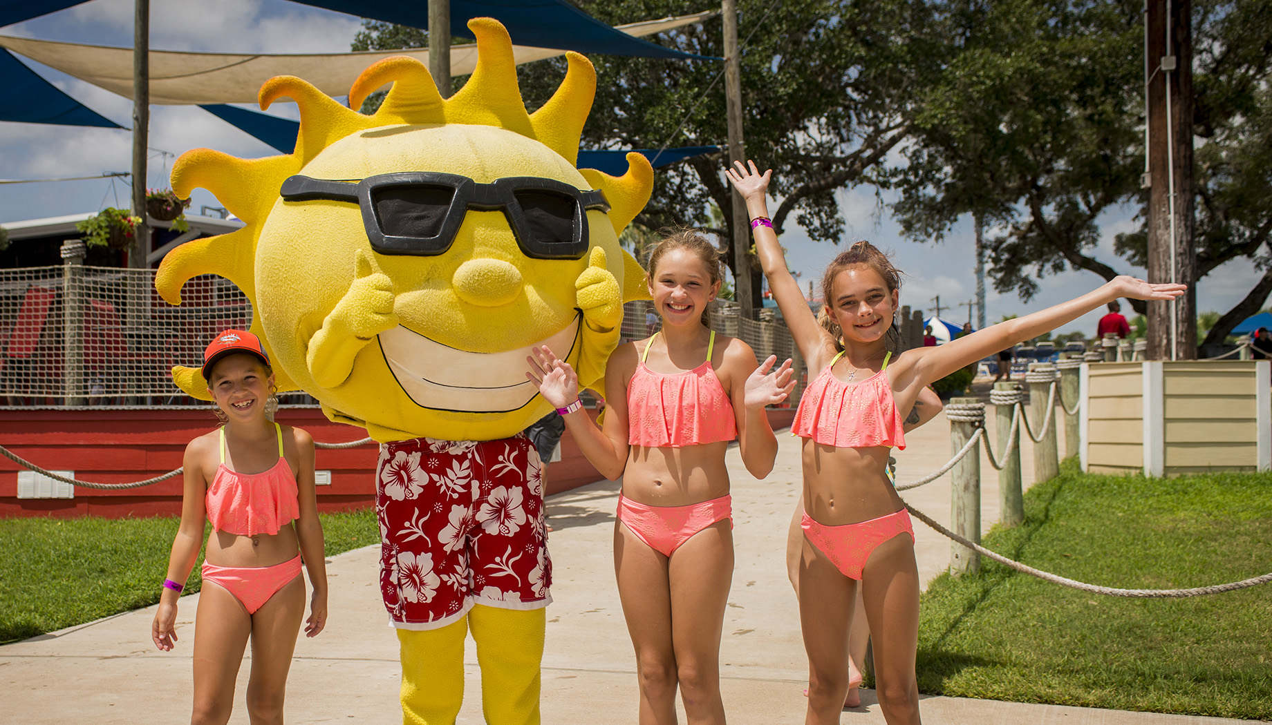 BLOG Three children in pink swimsuits pose with a person in a large sun mascot costume dressed in a red Hawaiian shirt with sunglasses, under sunny outdoor conditions.
