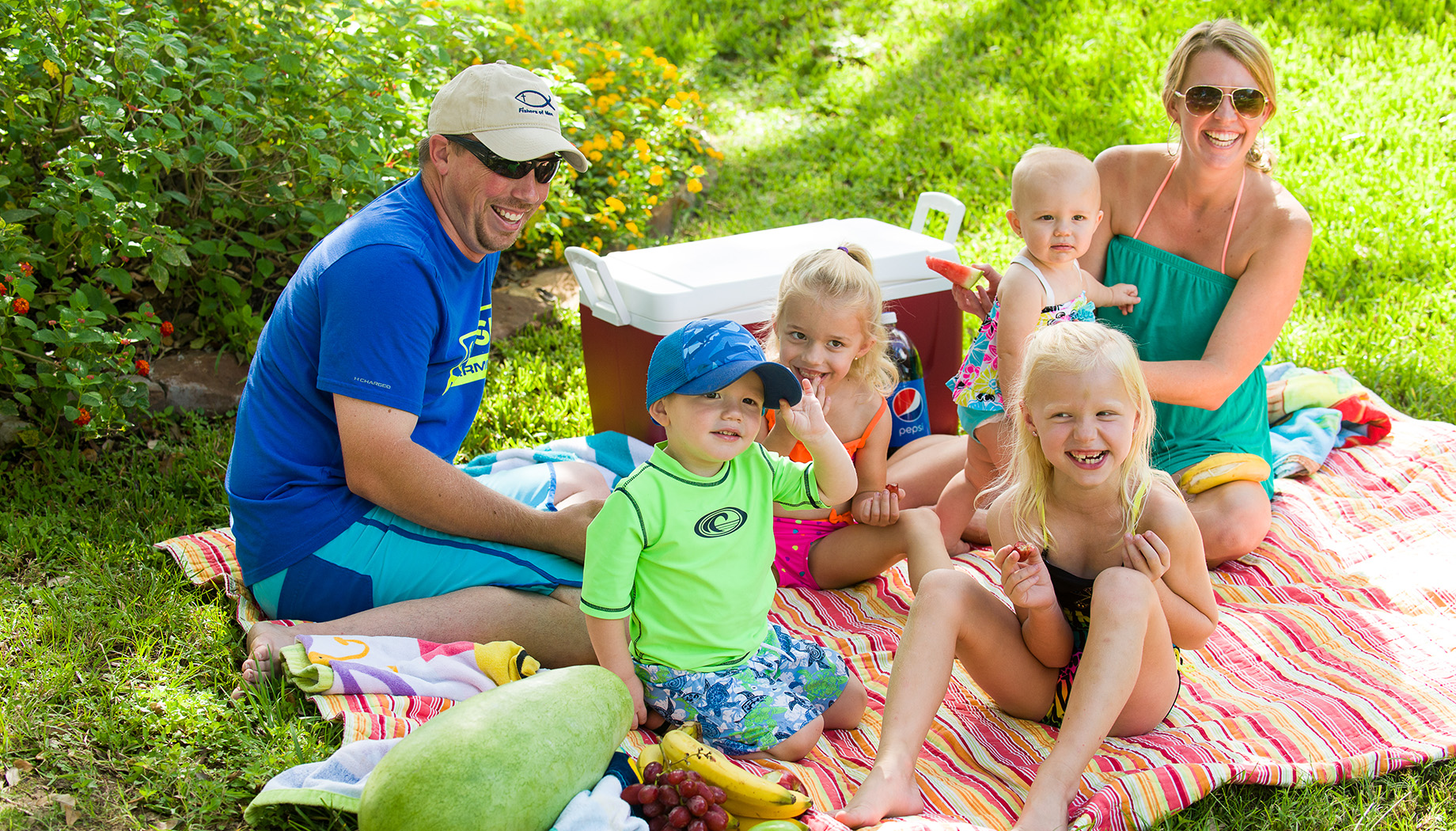 Bloggggg A family enjoys a picnic on a colorful blanket. Two adults and four children are sitting on the grass with a cooler and various fruits.