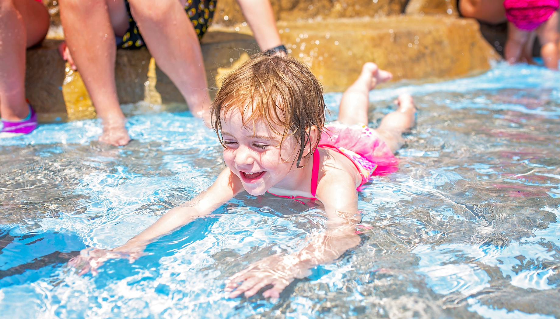 BLOG A young child in a pink swimsuit is smiling while lying on their stomach in shallow water at a pool, with other people sitting on the edge in the background.