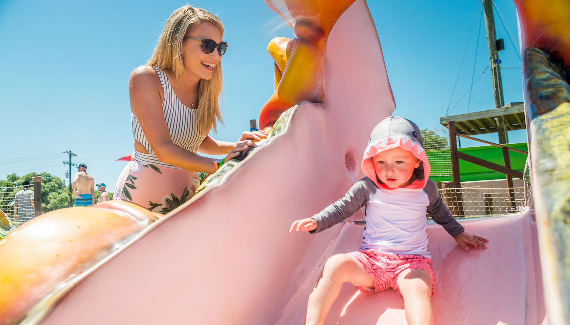 BLOG An adult assists a child riding down a colorful slide on a sunny day. The child wears a hooded shirt and shorts, and the adult is wearing sunglasses and a striped top.