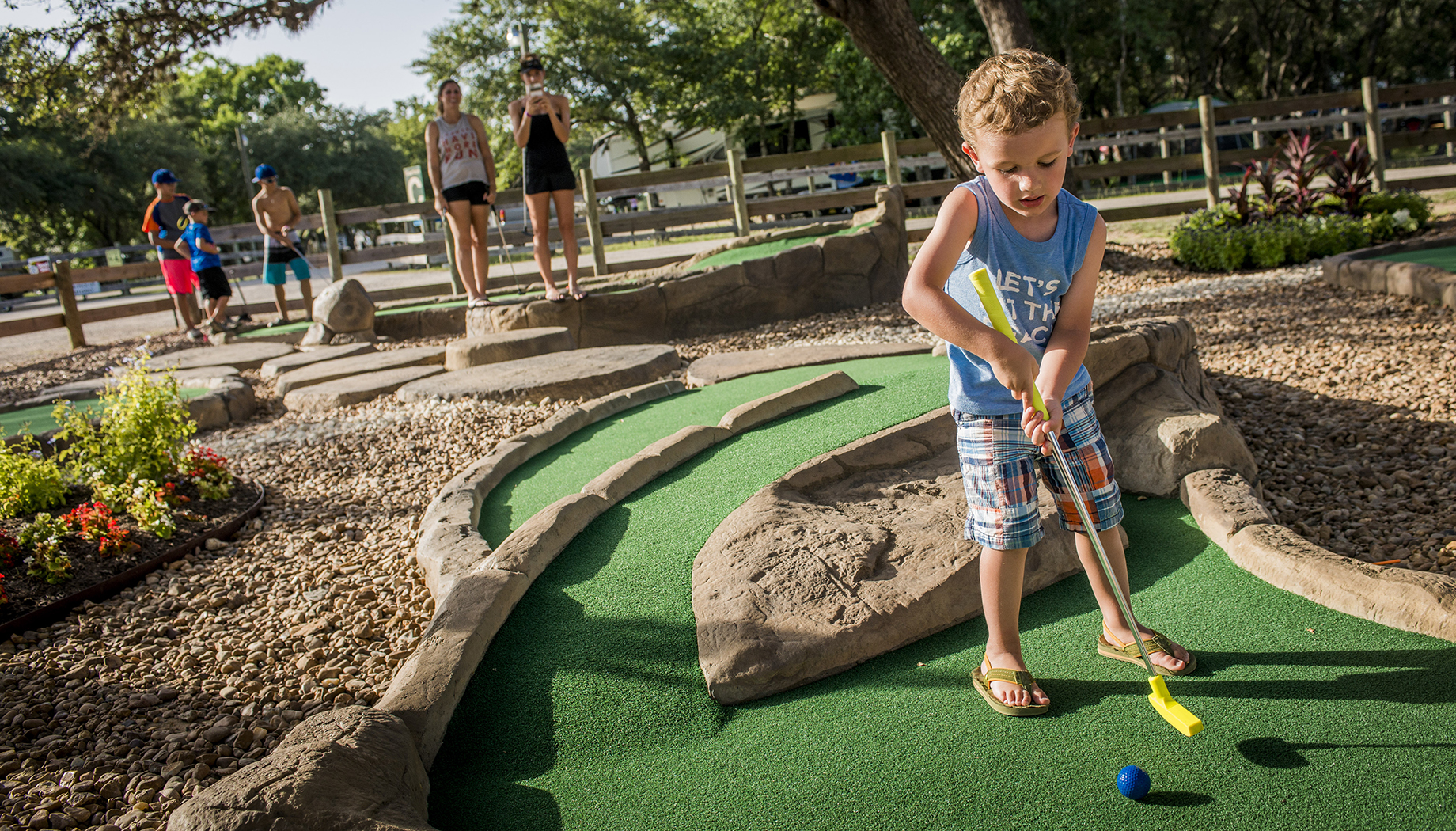BLOG A young boy wearing plaid shorts and a blue tank top plays mini golf on a course with rocks, as other people watch in the background.