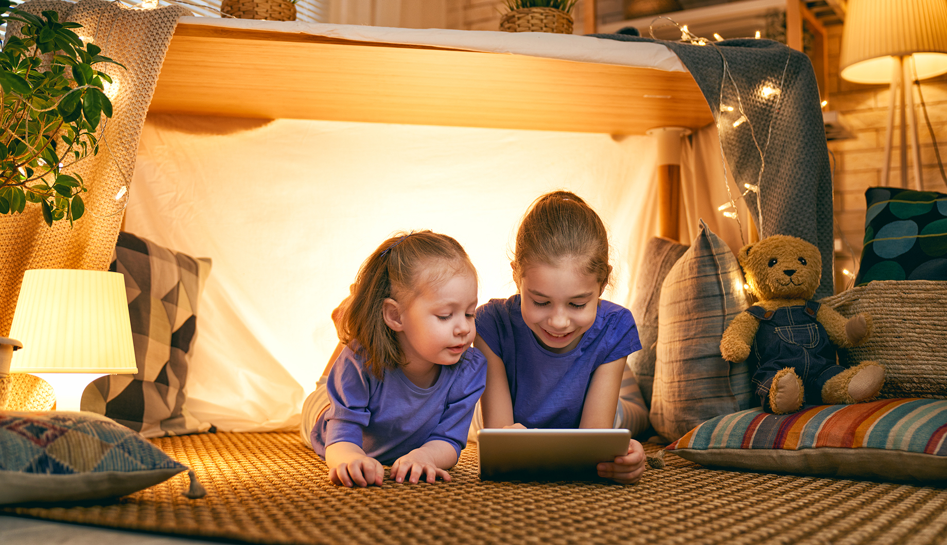 children are playing with tablet in tent Two children wearing blue shirts are lying on the floor inside a cozy blanket fort, illuminated by string lights, looking at a tablet.