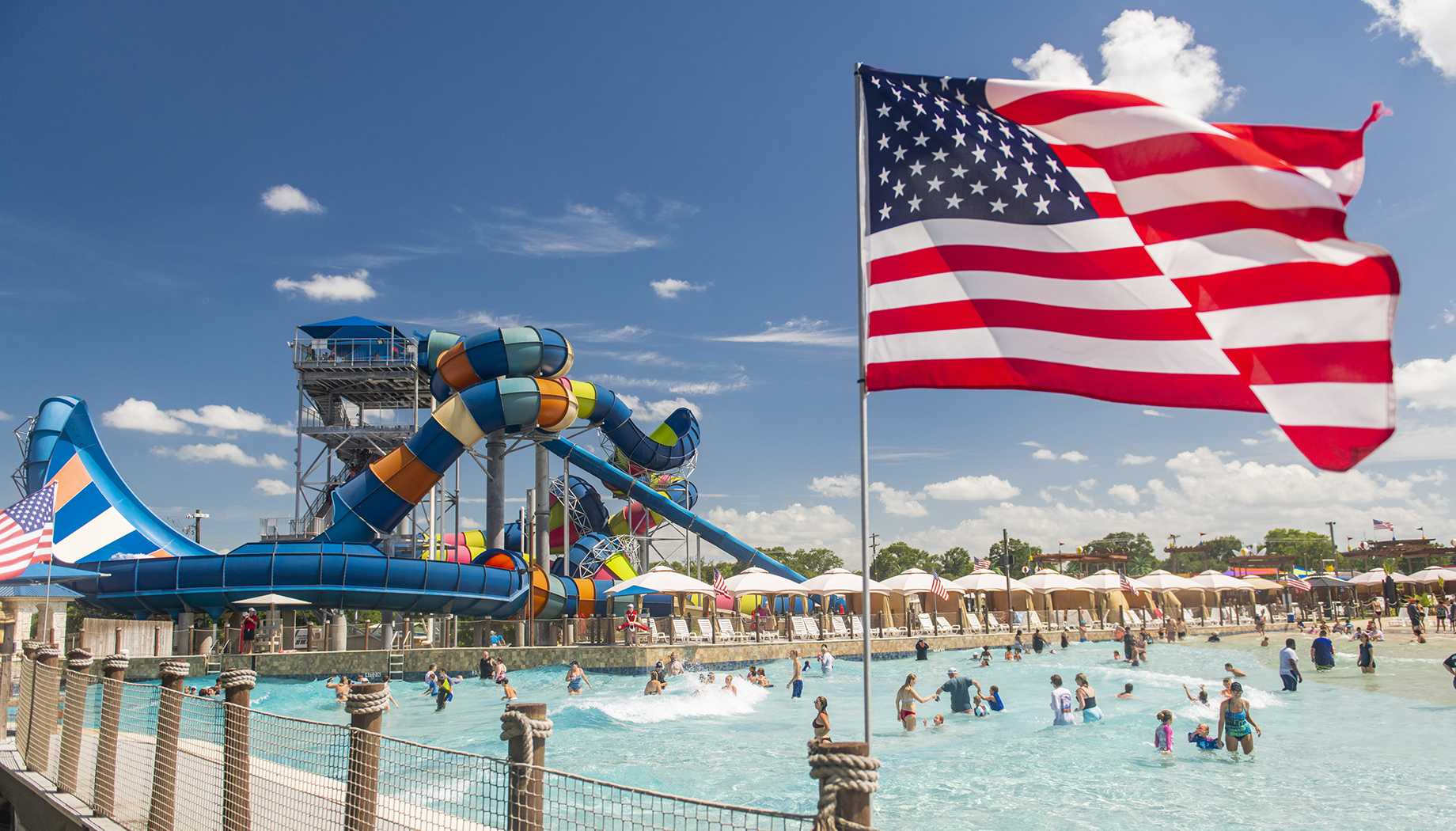 People enjoying a day at a water park with several slides and a wave pool. An American flag waves in the foreground under a blue sky.