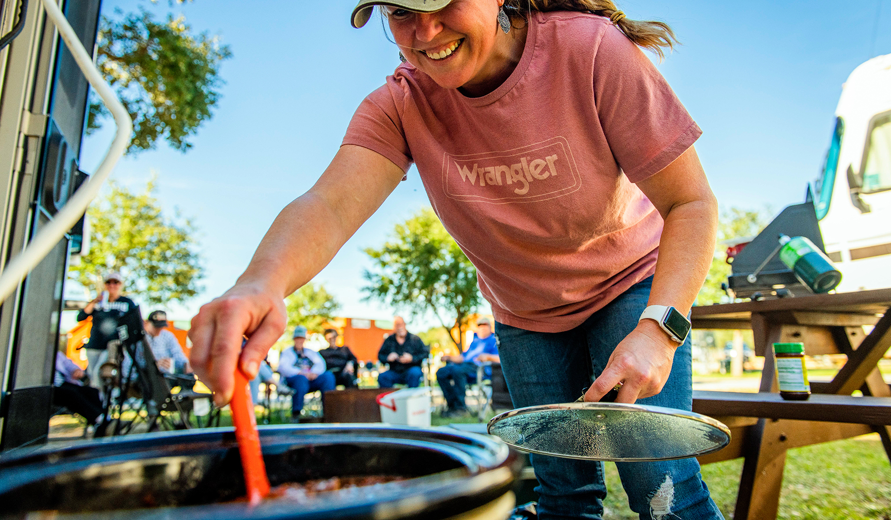 BLOG Woman stirring chili in a crockpot with an RV and people in the background.